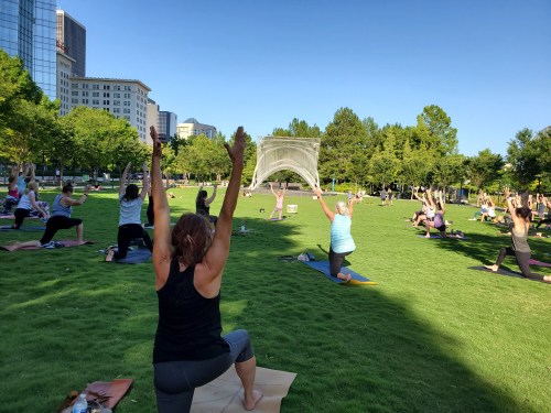 A group of people practicing yoga outdoors in a park, with some in seated poses and others stretching with arms raised.