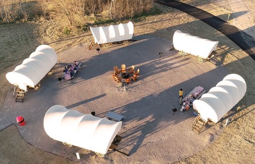 Aerial view of covered wagons arranged in a circle around a central fire pit, with people gathered nearby.