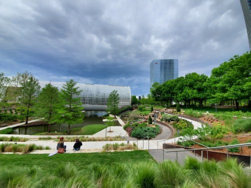 A serene garden with a pond, pathways, and modern buildings under a cloudy sky, featuring lush greenery and flowers.