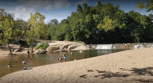 A sandy riverbank with people swimming and playing in the water, surrounded by trees and a small waterfall.