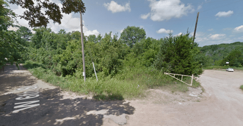 A dirt road intersects with a grassy area, surrounded by trees and power lines under a blue sky with clouds.