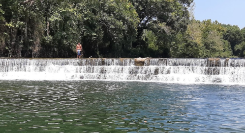 A serene waterfall cascades over rocks into a calm green pool, surrounded by lush trees and a clear blue sky.