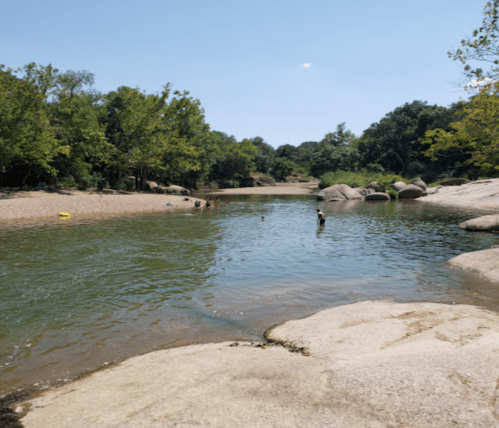 A serene river scene with people swimming, surrounded by lush greenery and large rocks under a clear blue sky.