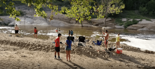 A sandy riverbank scene with people fishing, relaxing, and enjoying the water on a sunny day.