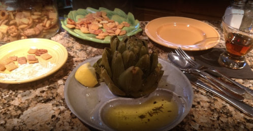 A cooked artichoke with lemon and olive oil, surrounded by plates of snacks and a glass of tea on a granite countertop.