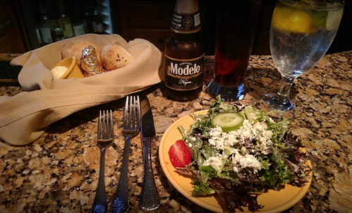 A plate of salad with cucumbers and tomatoes, accompanied by drinks and rolls on a granite table.