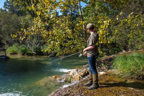 A person in camouflage fishing by a river, surrounded by trees and greenery on a sunny day.