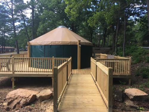 A wooden walkway leads to a green and tan yurt surrounded by trees and rocks.