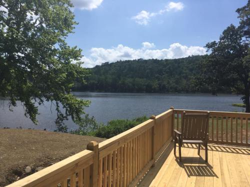 A wooden deck overlooks a calm lake surrounded by trees and hills under a blue sky with fluffy clouds.