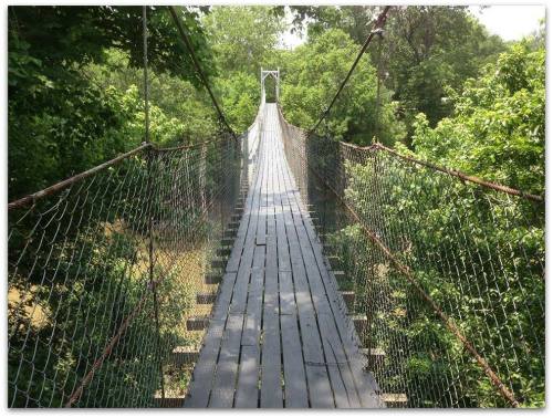 A wooden suspension bridge stretches over a lush green landscape, surrounded by trees on both sides.