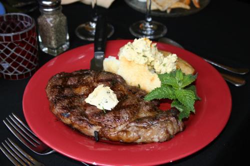 A juicy steak topped with herb butter, served with a side of cornbread and garnished with mint on a red plate.