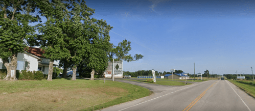 A rural road with a sign for a business, flanked by trees and a house on the left, under a clear blue sky.