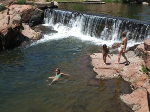 Three children play near a waterfall, with one wading in the water and two standing on the rocks by the shore.