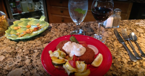 A plate of fried food with vegetables, garnished with sauce, alongside glasses of water and red wine on a granite countertop.
