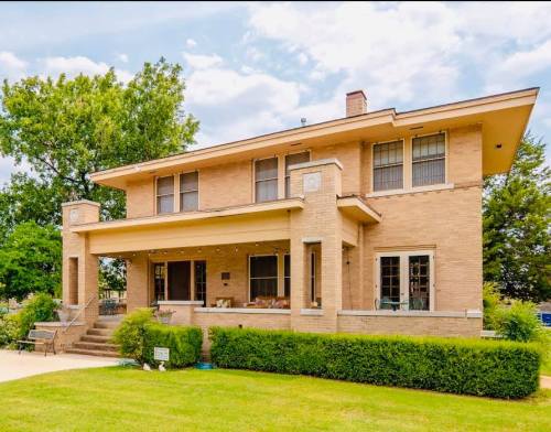 A two-story brick house with a large porch, surrounded by green grass and trees under a partly cloudy sky.