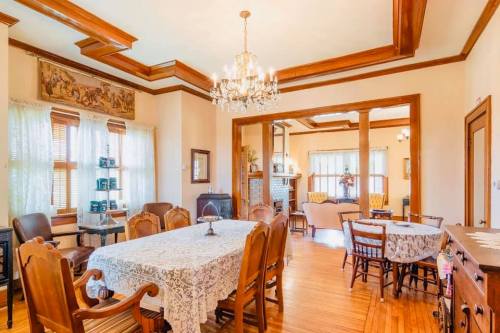 A spacious dining room with a large table, elegant chandelier, and wooden accents, featuring a cozy living area in the background.