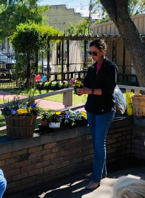A woman in a black jacket plants flowers in a pot, surrounded by blooming plants in a sunny outdoor setting.