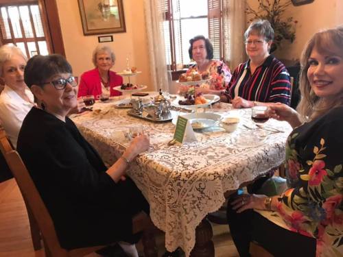 A group of six women enjoying tea and snacks around a beautifully set table with lace decor.