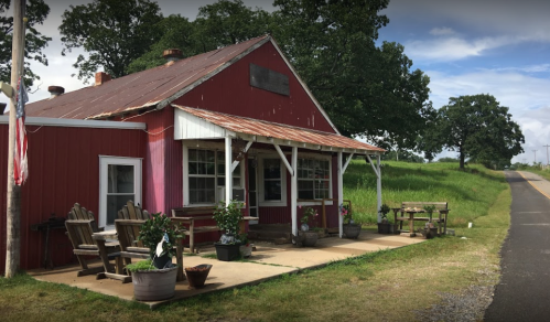 A rustic red building with a metal roof, surrounded by greenery, featuring a porch and outdoor seating.
