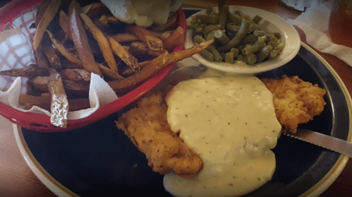 A plate of fried chicken topped with creamy gravy, served with green beans and a basket of crispy fries.