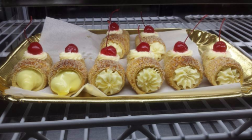 A tray of cream-filled pastries topped with whipped cream and cherries, displayed in a refrigerated case.