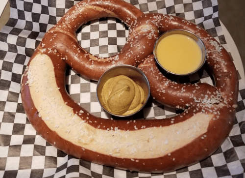 A large, soft pretzel with coarse salt, served with mustard and cheese dip in metal bowls on a checkered paper.