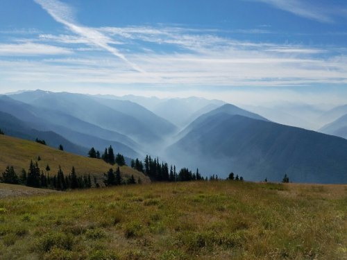 A panoramic view of misty mountains under a blue sky, with grassy hills in the foreground and layers of peaks in the distance.