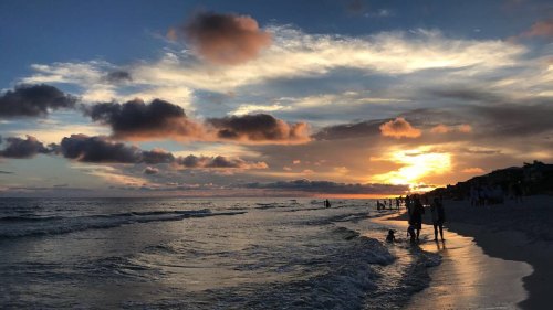 Silhouettes of people walking along a beach at sunset, with colorful clouds reflecting on the water.