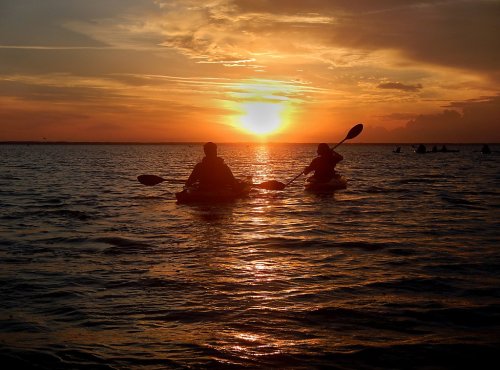Two kayakers paddle on a calm lake at sunset, with vibrant orange and yellow hues reflecting on the water.