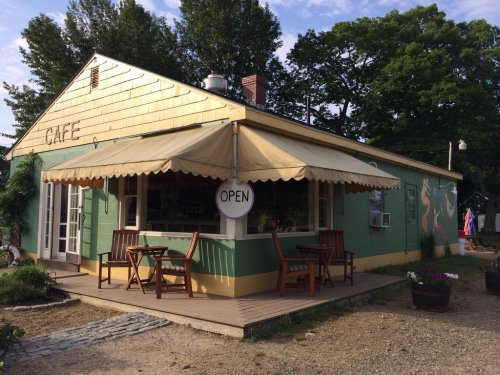 A charming café with a green exterior, open sign, and outdoor seating under a canopy, surrounded by trees.