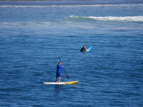 A person in a blue wetsuit paddleboards on calm water, while another person in a black wetsuit surfs nearby.
