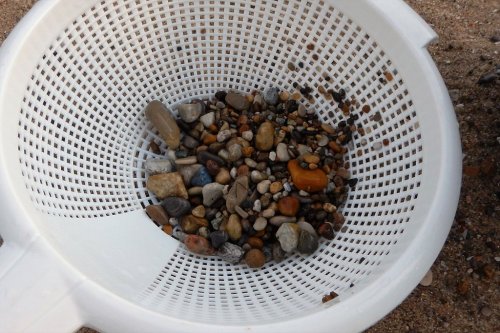 A white colander filled with various small stones and pebbles on a sandy surface.