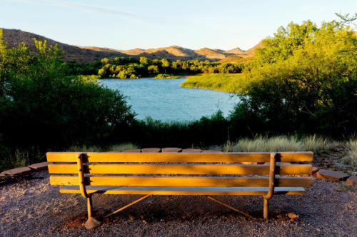 A wooden bench overlooks a serene lake surrounded by lush greenery and mountains under a clear sky.