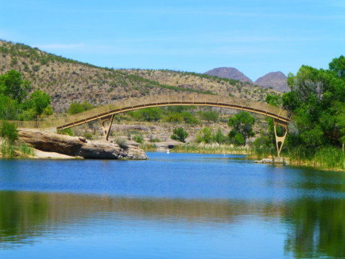 A wooden arch bridge spans a calm river, surrounded by lush greenery and rolling hills under a clear blue sky.