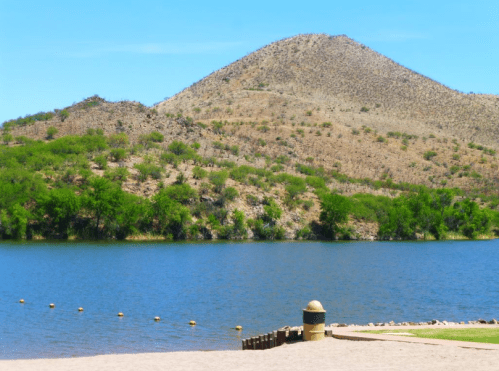A serene lake surrounded by green trees and a sandy shore, with a mountain in the background under a clear blue sky.