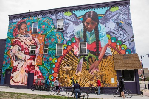 Colorful mural on a building featuring two women, one in traditional dress and another working in a field, with bicycles nearby.