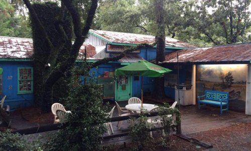 A colorful, rustic building surrounded by trees, featuring outdoor seating and a mural on the wall.