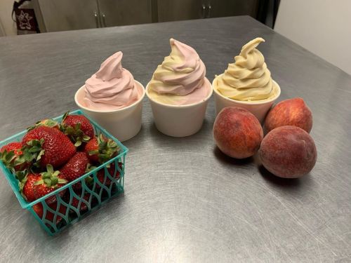 Three cups of soft serve ice cream in pink and cream colors, alongside a basket of strawberries and two peaches.