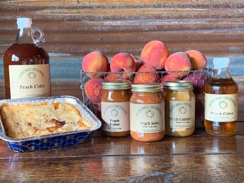 A basket of peaches next to jars of peach products and a dish of peach cobbler on a wooden table.