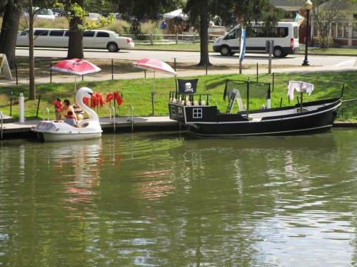 A swan-shaped float and a pirate-themed boat docked by a green park with umbrellas and trees in the background.