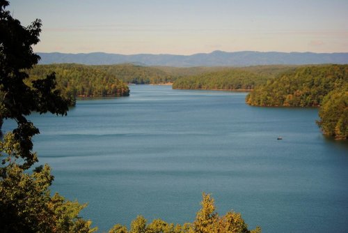A serene lake surrounded by lush green hills under a clear blue sky, with distant mountains in the background.