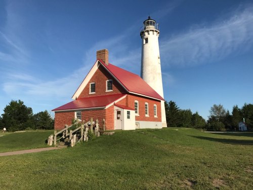 A tall white lighthouse beside a red-roofed brick building, set against a clear blue sky and green grass.