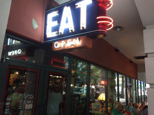 A restaurant entrance with a bright "EAT" sign and "OPEN" neon lights, featuring large windows and outdoor seating.