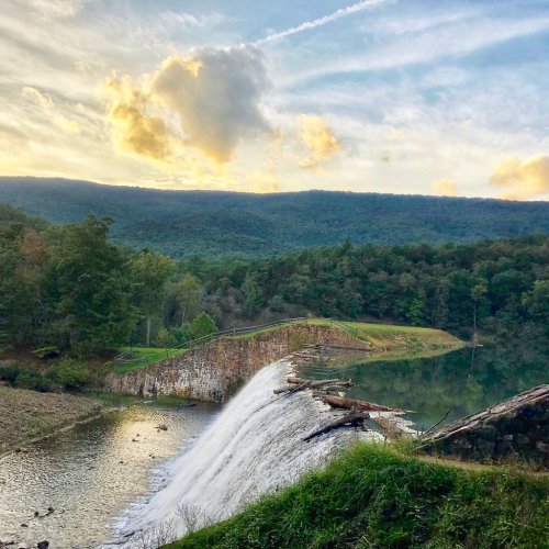 A serene landscape featuring a waterfall cascading over a dam, surrounded by lush greenery and a cloudy sky.