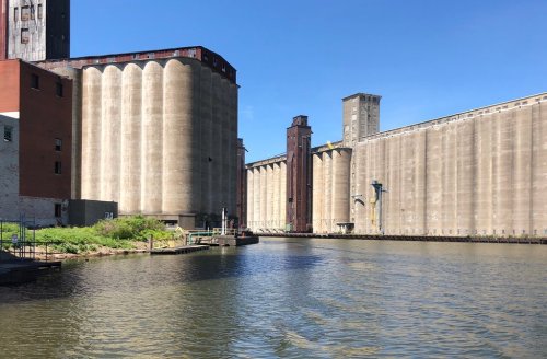 Industrial buildings along a riverbank under a clear blue sky, with silos and a calm water surface in the foreground.