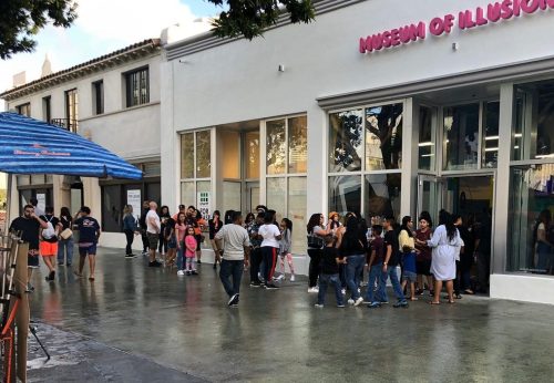 A crowd of people gathers outside the Museum of Illusions, with a blue umbrella and shops in the background.