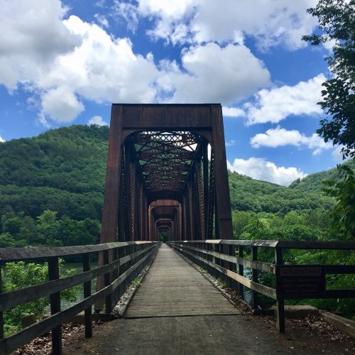 A rusty train bridge spans a lush green valley under a blue sky with fluffy clouds.