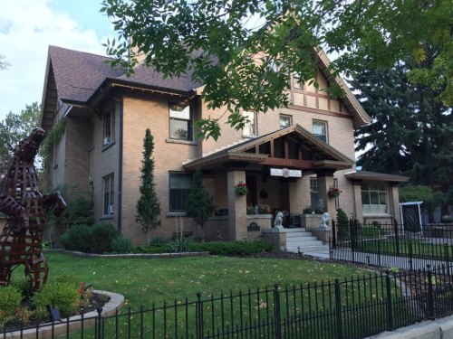 A large, two-story brick house with a gabled roof, surrounded by greenery and a decorative fence.