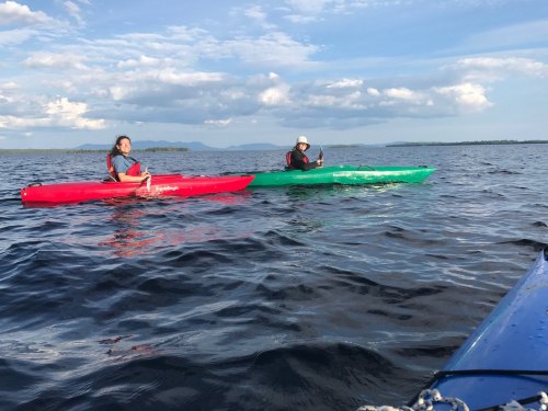 Two people kayaking on a calm lake, one in a red kayak and the other in a green kayak, with mountains in the background.
