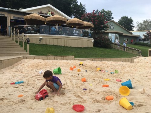 A child plays with a red toy car in a sandy area filled with colorful plastic toys, with a restaurant in the background.